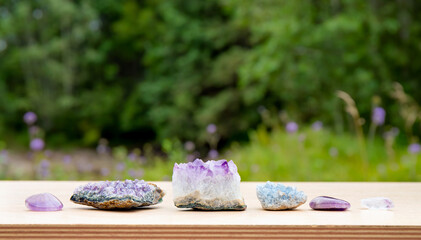 Different raw natural crystal geode clusters, amethyst, celestite, quartz on wood table with blurred wild green nature on background.