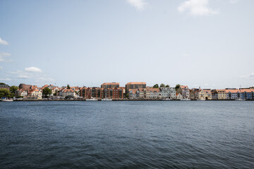 panoramic view of a picturesque waterfront town with colorful historic buildings lining the shore, reflected in calm water beneath a clear, open sky. Denmark