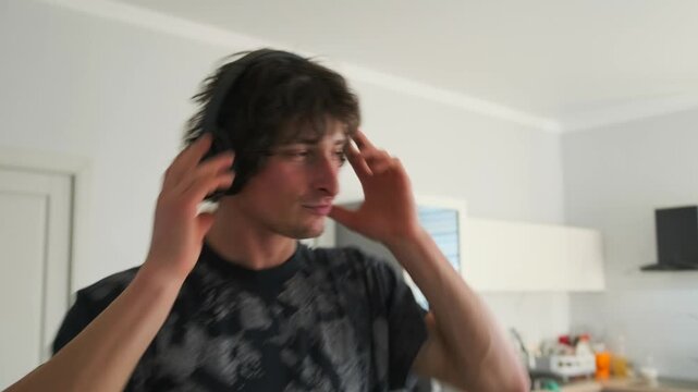 A young man in a black T-shirt and headphones dances in the kitchen to