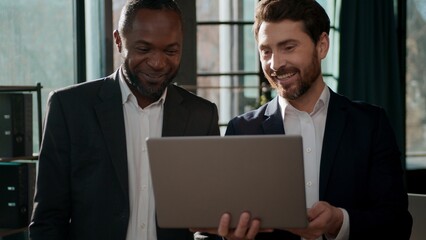 Two excited business team men businessmen looking laptop screen happy communicate in office discussing good idea excellent result project success rejoice Caucasian African American coworkers teamwork