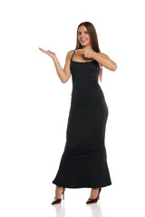 Studio shot of a smiling woman with dark hair wearing a long black dress and high heels, standing and pointing while presenting with open palm, full body, on a white background