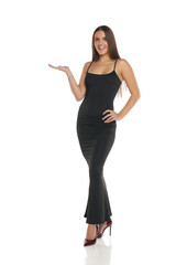 Studio shot of a smiling woman with dark hair wearing a long black dress and high heels, standing and presenting with open palm, full body, on a white background