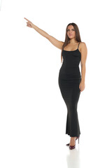 Studio shot of a woman with dark hair wearing a long black dress and high heels, standing and pointing to the side, full body, on a white background