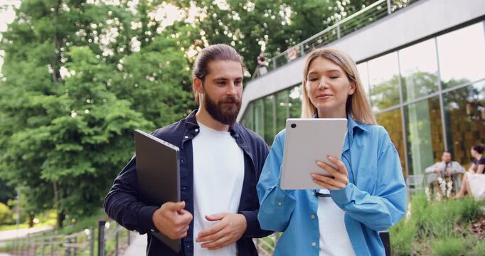 Young woman holding tablet and business partner looking at the screen together, engaged in a focused discussion about work processes during an outdoor business meeting