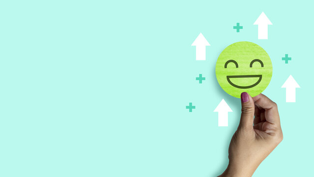 Woman hand holding a green smiling face paper circle with white arrows and a green plus sign around, on a light green background. Concept of Mental Health