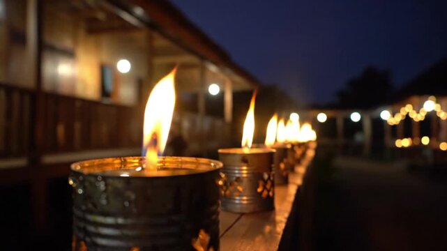 A closeup of a vibrant flame on a metal container with a line of similar glowing lamps extending into the blurred background set at dusk