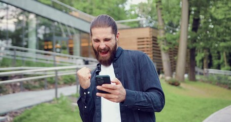 Surprised Caucasian man standing in a city park, using a smartphone and reacting emotionally to unexpected news. Concept of online communication and technology.