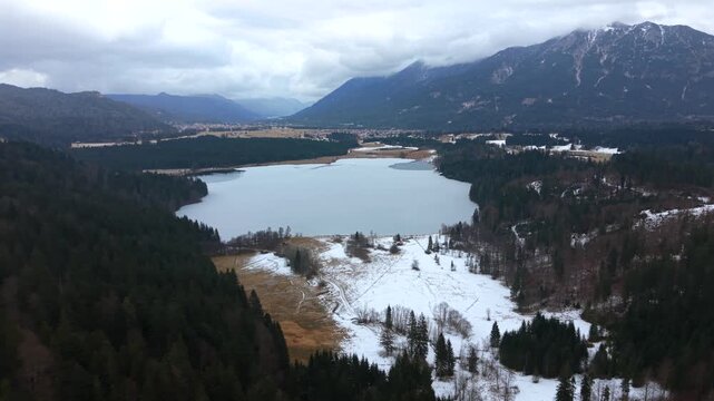 Cinematic aerial panorama of Lake Barmsee in Bavaria, Germany. Winter drone shot reveals frozen lake surface, surrounding forest and alpine meadows near the Karwendel mountains.
