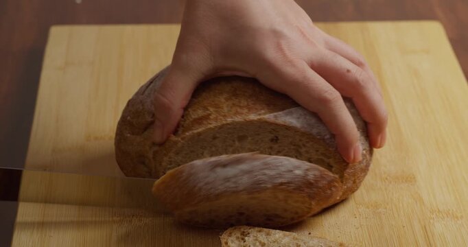 Close-up of cutting crusty multigrain sourdough bread in a home kitchen. Making fresh toast for breakfast. Hands slicing fresh French wholegrain bread on a table. Preparing crispy a homemade breakfast