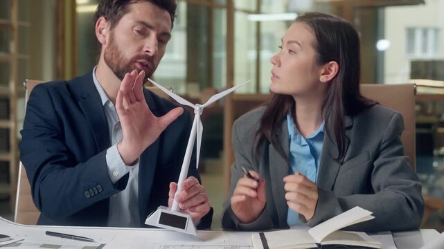 Multiracial teamwork Korean woman Caucasian man examine wind turbine model office desk collaboration sustainable energy solution brainstorming green tech clean prototype strategy business blueprint