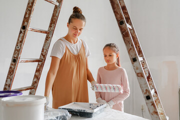 A mother guides her daughter as they paint a wall in their home, sharing a special moment during their weekend renovation project