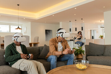 African American men playing with white VR headsets and controllers on gray sofa in living room