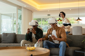Male friends playing VR games on sofa in living room, wearing white headsets, holding controllers