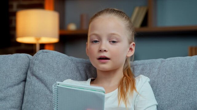 Caucasian little child girl kid daughter reading copybook talking looking at camera at home video call e-learning read text holding textbook on sofa online conference communication learning education