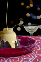 Christmas pudding with custard being poured on it and a glass of champagne on a black background with Christmas lights