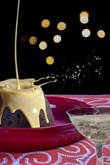Christmas pudding with custard being poured on it and a glass of champagne on a black background with Christmas lights
