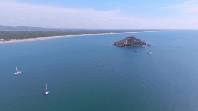 Vista a&eacute;rea de la Isla de la Piedra en Mazatl&aacute;n, M&eacute;xico, que muestra tranquilas aguas azules del oc&eacute;ano, peque&ntilde;os veleros anclados en alta mar y una costa arenosa bajo un cielo despejado y soleado. Ap