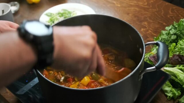 A male hand stirs vegetable stew in a pot with a wooden spatula as pep