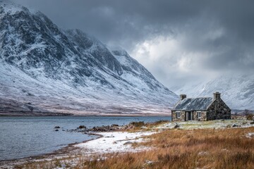 Stone cottage beside a lake nestled at base of snowy mountains under a cloudy sky