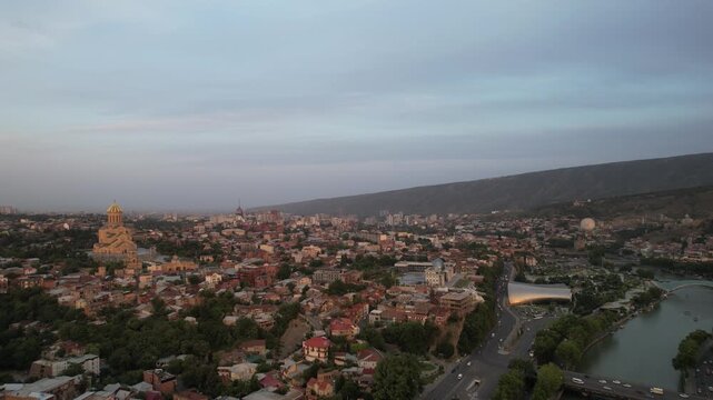 Aerial view of the Avlabari district in Tbilisi at dusk, flying above the Mtkvari Kura River. Warm sunset light and calm evening atmosphere. High-quality 4K footage.