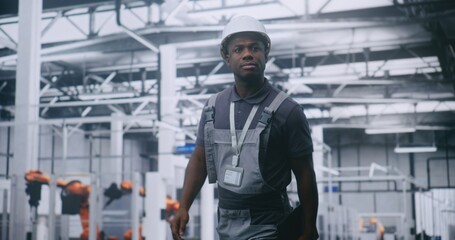African American Male Factory Supervisor Walking Across High Tech Manufacturing Facility, Using Tablet Check Production Data, Assess Machinery Performance and Oversee Automated Industrial Operations.