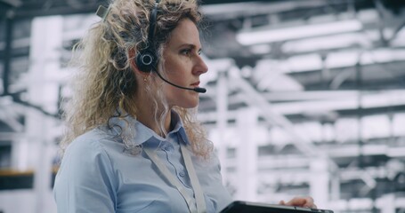 Close Up of Female Factory Engineer Wearing Communication Headset and Actively Engaging With Digital Tablet. Real Time Coordination, Remote Operations, and Digital Communication on Industrial Floor.