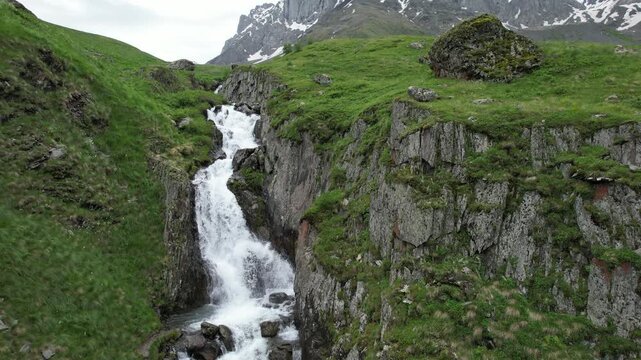Aerial drone view approaching a waterfall along a mountain trail near Juta, Georgia, with the Chaukhi Pass and dramatic alpine peaks in the background. High-quality 4K footage.