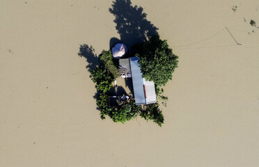 A top-down aerial view of a lone house and trees surrounded by floodwaters, capturing isolation, vulnerability, and the severe impact of flooding on rural livelihoods