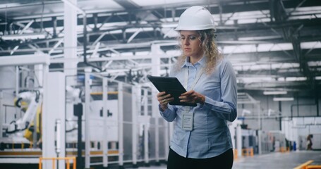 Professional Female Engineer in Hard Hat Checks Production Data on Tablet While Walking Through Modern Automated Factory. Concept of Smart Manufacturing, Industry 4.0, Digital Control and Efficiency.