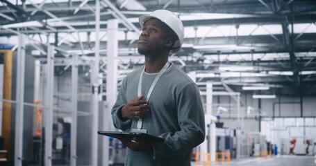 Adult Male Engineer Wearing Safety Helmet, Holding Tablet, and Looking Away to Survey Production Floor. Vast Industrial Interior Emphasizes Strategic Management and Critical On Site Planning.