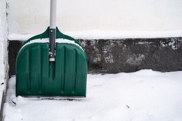 Green snow shovel standing against a wall on a snowy ground. Winter tool used for clearing snow outdoors. Seasonal weather concept with equipment for snow removal.