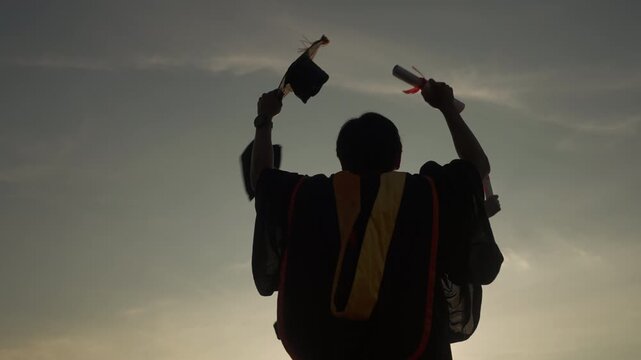 Two graduates in academic gowns raising cap and diploma under sunrise, celebrating education success, freedom, and a new chapter in life.