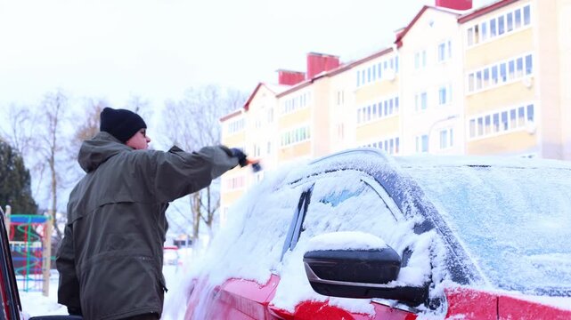 A young Caucasian man with dark hair clears snow from a red car in a winter setting. Apartment buildings are visible in the background.