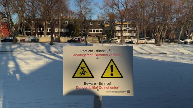 Warning sign in Muenchen, Germany, cautioning about thin ice on the Nymphenburg Canal. Text reads Beware thin ice. Danger to life. in German and English on a snowy winter day in Bavaria