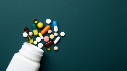 Colorful medicinal pills and capsules spill from a white plastic bottle onto a dark teal surface