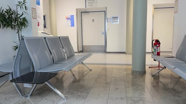 Interior view of an empty waiting area in a German hospital. Features modern metal benches for patients, a clean tiled floor, and a door marked with number 8. Reflects healthcare in Germany