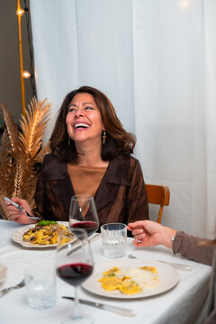 Vertical lifestyle portrait of joyful woman laughing during cozy dinner with pasta and red wine