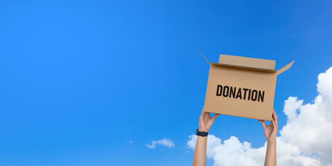 Person raising a cardboard donation box toward a clear blue sky.