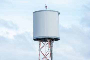 White Industrial Water Tank on Metal Lattice Tower Against Bright Sky, Minimalist View of a Public Utility Water Tower and Communication Mast