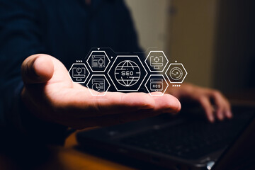A hand holding digital icons representing SEO and online marketing concepts, with a laptop visible...