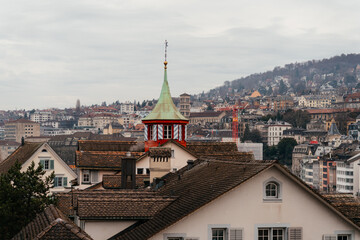 A panoramic cityscape of Zurich featuring historic church towers and rooftops under a cloudy sky. The image captures the charm of Switzerland old town with mountains in the background.