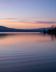Fototapeta premium Serene lakeside landscape reflecting a vibrant sunset over distant mountains during dusk