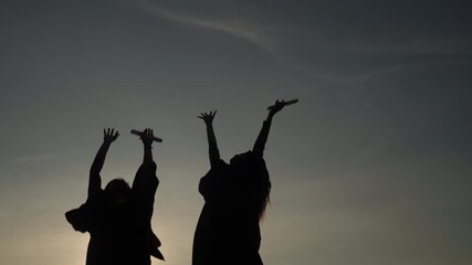 Silhouette of two graduates throwing graduation cap in the air and holding diplomas during sunset, celebrating freedom and academic success.