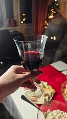 Hand holding glass of red wine above festive table with food and blurred christmas lights in background. This image represents celebration holiday atmosphere and cozy home gathering moment