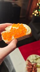 Bread slice with butter and red caviar held in hand over festive table with warm lights in background. This image represents luxury holiday tradition and intimate celebration moment