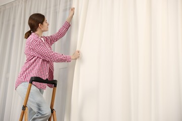 Woman with ladder hanging up window curtains at home, low angle view. Space for text © New Africa
