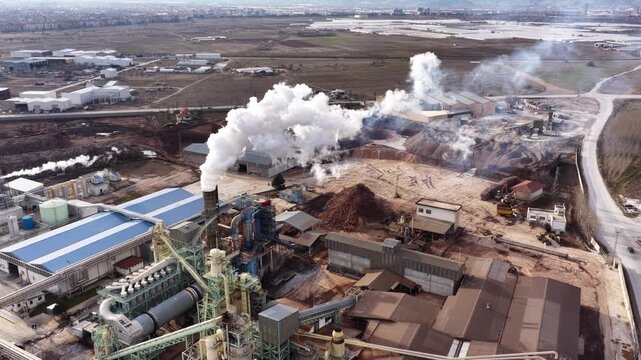 White smoke rising from the particleboard and MDF factory. Aerial drone view of the industrial facility's raw material yard, production lines, and surrounding residential areas.