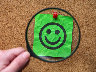 A person with a magnifying glass in his hand magnifying a green note paper with a happy face on it. Close up.