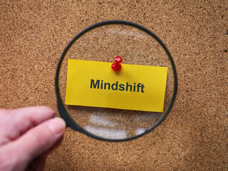 A person with a magnifying glass in his hand magnifying a yellow piece of paper with the word Mindshift on it. Close up.