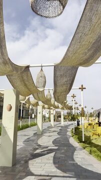 Outdoor seating area at Suq Al-Uyun Market featuring a paved pedestrian walkway shaded by flowing fabric canopies and yellow wooden crate furniture against a scenic mountain range under a cloudy sky.